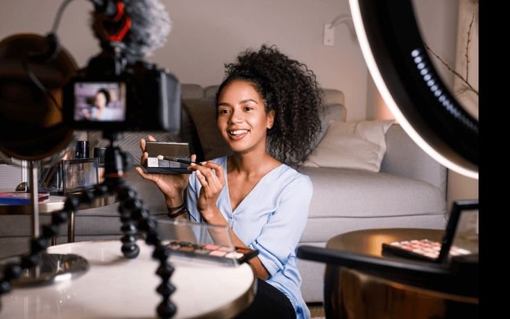 "Female beauty influencer sitting in front of a camera and ring light, demonstrating an eyeshadow palette during a makeup tutorial, with cosmetics arranged on the table."